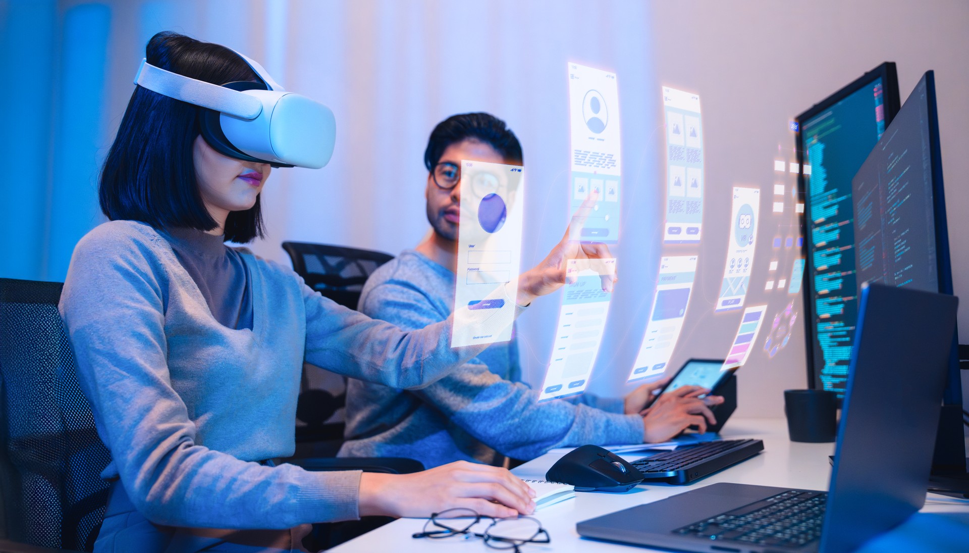 Asian software developer wearing a virtual reality headset works on a VR project, with a colleague focused on her computer